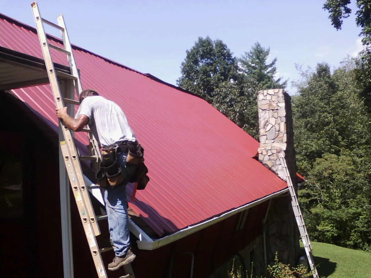 Roofer on a ladder beside a red metal roof and stone chimney — Soffit & Fascia Repair by Roofing Brothers in Orchard Park